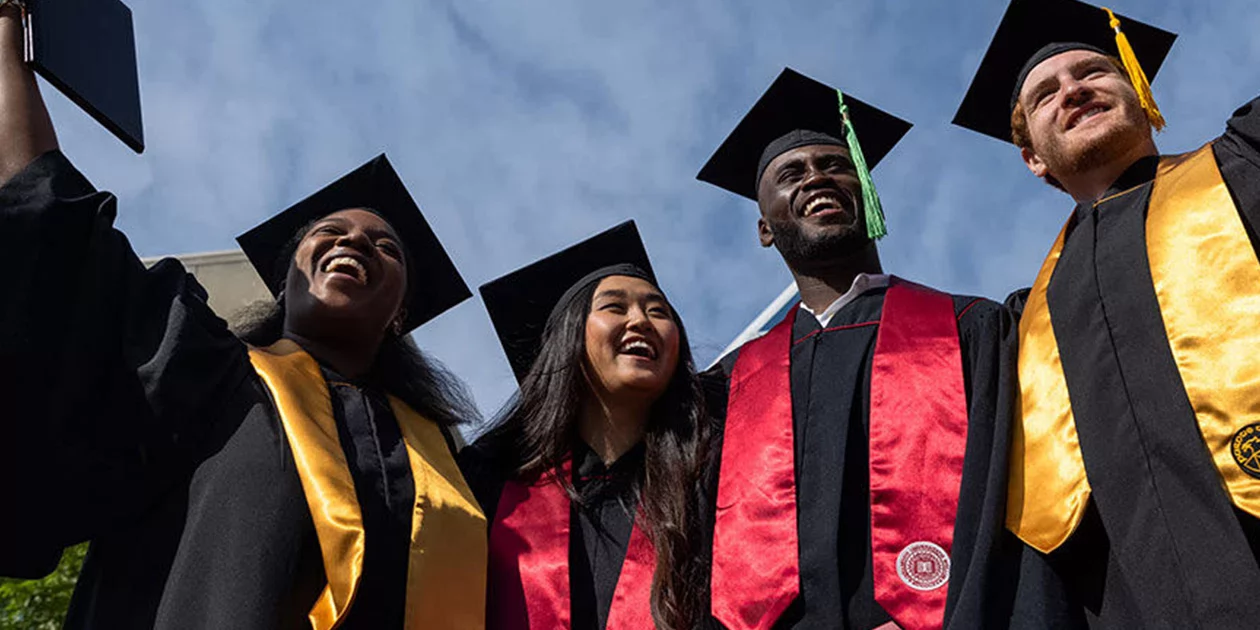 Group of IU Indianapolis first generation college students in caps and gowns smiling together outdoors under a blue sky during commencement.