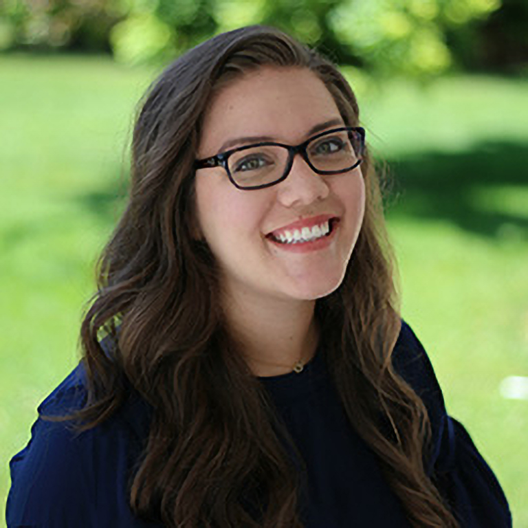 A young woman with long brown hair and glasses smiling outdoors with a blurred green background.